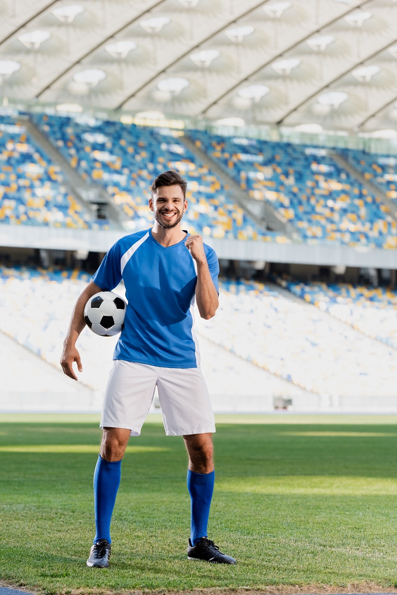 happy professional soccer player in blue and white uniform with ball showing yes gesture on football
